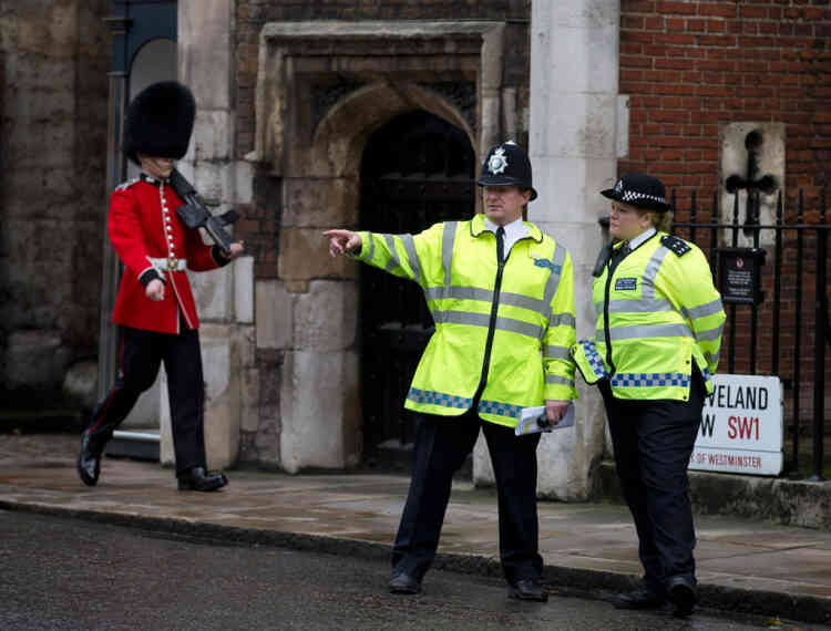 Police officers monitor the small crowd of media and royal fans outside St James's Palace, as a member of the Grenadier Guards marches past in London, Wednesday, Oct. 23, 2013. Prince William and his wife Kate have asked seven people to be godparents to their son, Prince George, who will be christened at a major royal family gathering Wednesday, palace officials said.  Queen Elizabeth II and her husband Prince Philip plan to attend the christening Wednesday at the Chapel Royal at St. James's Palace, along with Prince Charles, his wife Camilla, Prince Harry and other royals. (AP Photo/Alastair Grant)