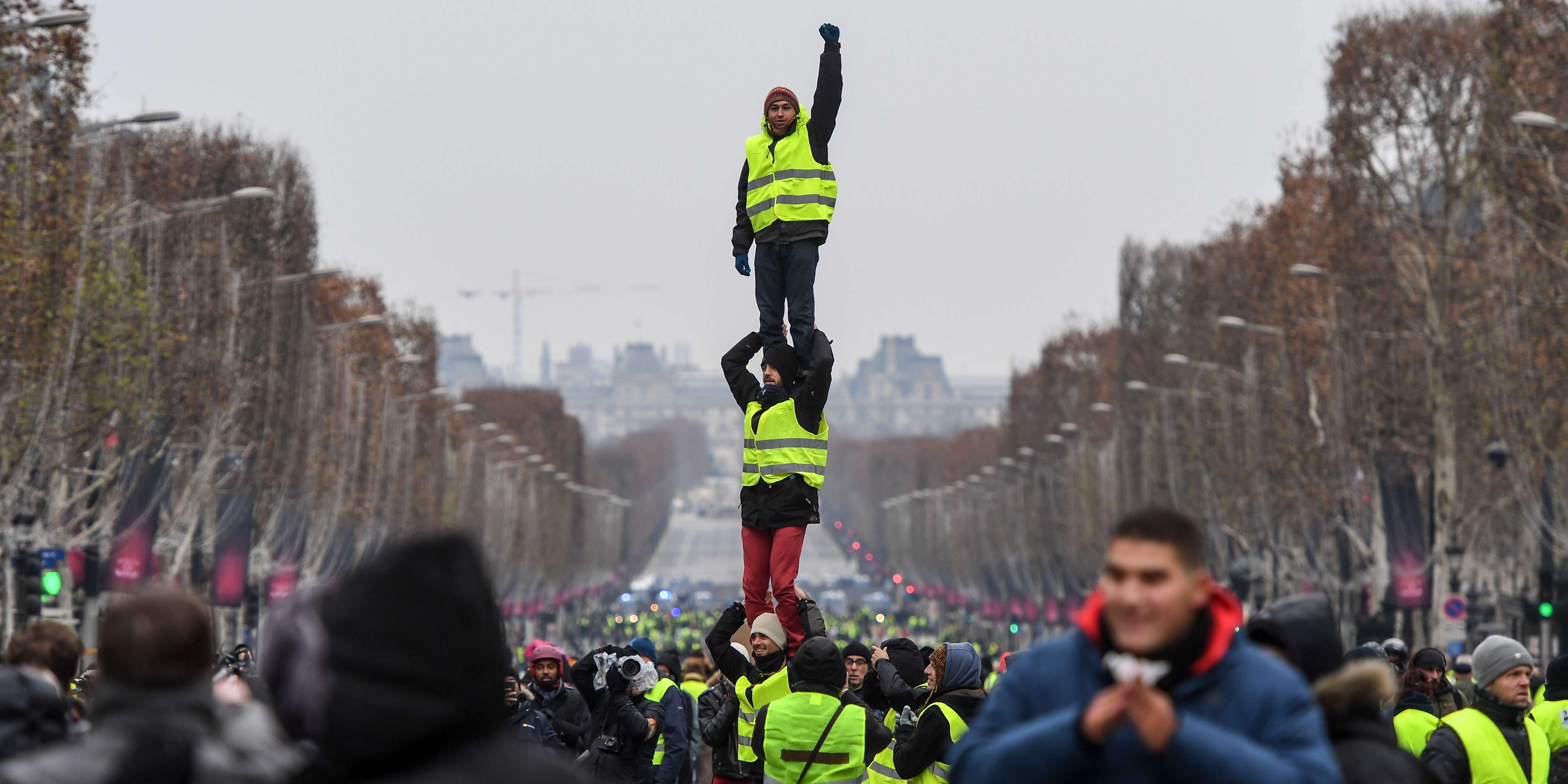 Protesters wearing yellow vests (gilets jaunes) do a human pyramide as they demonstrate against rising costs of living they blame on high taxes in Paris, on December 15, 2018. The "Yellow Vests" (Gilets Jaunes) movement in France originally started as a protest about planned fuel hikes but has morphed into a mass protest against President's policies and top-down style of governing. / AFP / Christophe ARCHAMBAULT