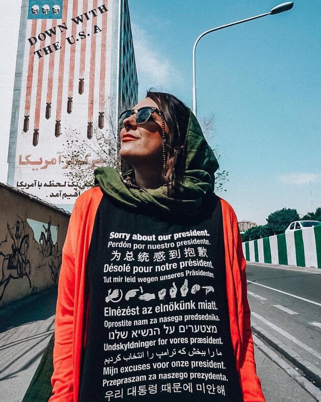 A Téhéran, en août 2018, une touriste américaine revêtue d’un tee-shirt portant des phrases d’excuse liées au comportement du président Trump. En arrière-plan, des slogans anti-américains sur une façade d’immeuble.