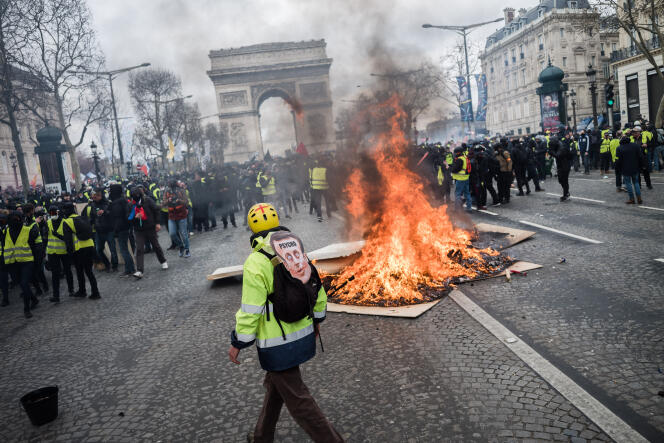A Paris, le 16 mars 2019, lors de l’acte XVIII du mouvement des « gilets jaunes ». Un manifestant portant un portrait du président de la Republique marche près d’un feu allumé au milieu des Champs-Elysees, non loin de la place de l’Etoile.