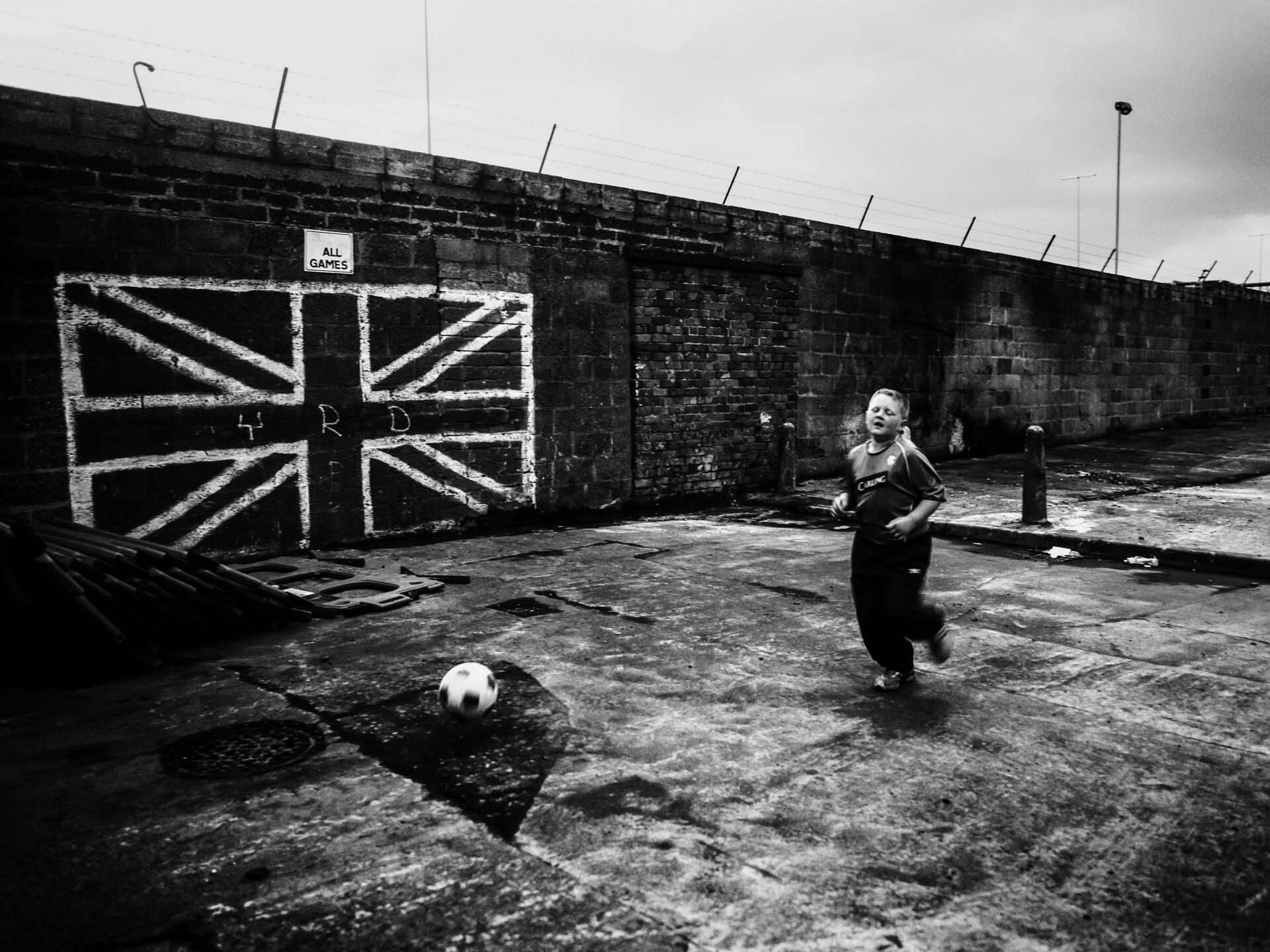 Un garçon joue au football devant le drapeau anglais peint sur le mur , dans le quartier de Shankill à Belfast. Extrait du livre « Wee Muckers – Youth of Belfast », de Toby Binder.