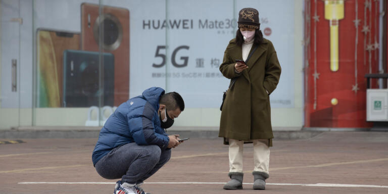 In this Sunday, March 8, 2020, photo, residents wearing masks look at their smartphones near an advertisement for 5G smartphones from Chinese tech giant Huawei in Beijing. Factories in China, struggling to reopen after the coronavirus shut down the economy, face a new threat from U.S. anti-disease controls that might disrupt the flow of microchips and other components they need. For most people, the new coronavirus causes only mild or moderate symptoms. For some it can cause more severe illness. (AP Photo/Ng Han Guan)