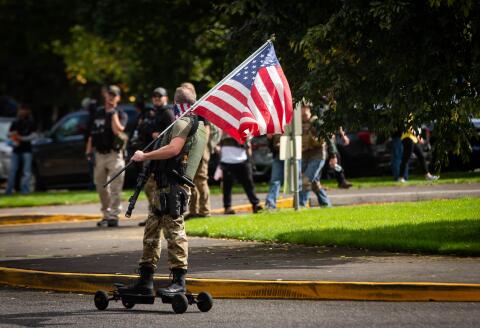 An armed atteendee skateboards waving an american flag as members of the Proud Boys and other similar groups attend a rally at Delta Park in Portland, Oregon on September 26, 2020. Far-right group "Proud Boys" members gather in Portland to show support to US president Donald Trump and to condemn violence that have been occurring for more than three months during "Black Lives Matter" and "Antifa" protests. / AFP / Maranie R. STAAB
