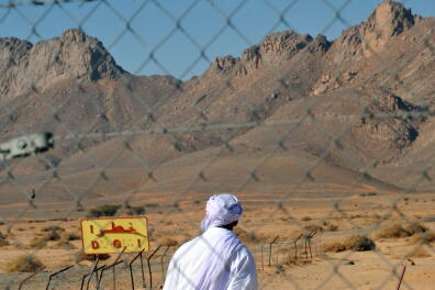An Algerian man is pictured through a fence as he looks at a danger sign at the entrance of the nuclear bomb test site of Tena Fila mountain at Ain Ekra in Tamanrasset 2000 km south of Algiers, on February 25, 2010. During an international conference on nuclear testing in Algiers on February 25 a series of propositions have been presented to efficiently combat the effects of the tests. In the irradiated Algerian desert children are still born with deformities that campaigners blame on the experimental blasts by the French during their occupation of Algeria in the 1960s. AFP PHOTO / FAYEZ NURELDINE (Photo by FAYEZ NURELDINE / AFP)