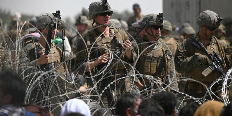 TOPSHOT - US soldiers stand guard behind barbed wire as Afghans sit on a roadside near the military part of the airport in Kabul on August 20, 2021, hoping to flee from the country after the Taliban's military takeover of Afghanistan. (Photo by Wakil KOHSAR / AFP)
