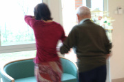 Inside a retirement home, a young woman supports an elderly man in the hall.