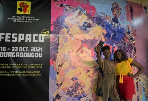 Women take a selfie next to a poster of the FESPACO headquarter during the 27th Pan-African film and television festival(FESPACO), Africa's biggest film festival in Ouagadougou, on October 20, 2021. - The films will be screened in various cinemas in Ouagadougou and there will also be screenings in ten open-air spaces. Of the 1,132 films entered, 17 feature-length fiction films have been selected for the official competition and are in the running for the 'Etalon d'Or du Yennenga', Fespaco's highest award. The directors came from 15 countries of the continent. (Photo by Issouf SANOGO / AFP)