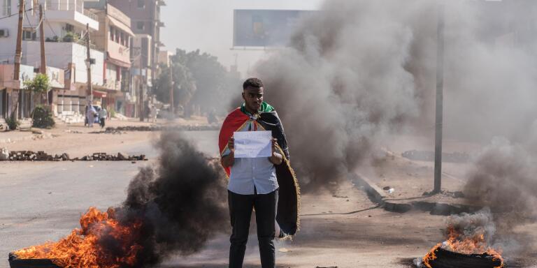 Un manifestant devant des barricades porte un drapeau soudanais sur le dos et tient une feuille où il est écrit 