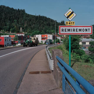 Le panneau d'entree de la ville avec au loin le rond-point ou se trouvent les commerces. 
Sur un rond-point a l'entree de Remiremont, deux modeles de boulangeries s'opposent. Remiremont, France, 18 juin 2022, pour Le Monde.