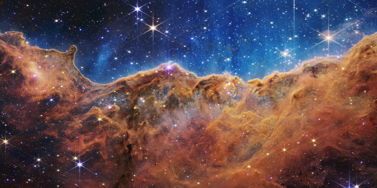 What looks much like craggy mountains on a moonlit evening is actually the edge of a nearby, young, star-forming region NGC 3324 in the Carina Nebula. Captured in infrared light by the Near-Infrared Camera (NIRCam) on NASA's James Webb Space Telescope, this image reveals previously obscured areas of star birth.Called the Cosmic Cliffs, the region is actually the edge of a gigantic, gaseous cavity within NGC 3324, roughly 7,600 light-years away. The cavernous area has been carved from the nebula by the intense ultraviolet radiation and stellar winds from extremely massive, hot, young stars located in the center of the bubble, above the area shown in this image. The high-energy radiation from these stars is sculpting the nebula's wall by slowly eroding it away.  NIRCam - with its crisp resolution and unparalleled sensitivity - unveils hundreds of previously hidden stars, and even numerous background galaxies. Several prominent features in this image are described below.-The 