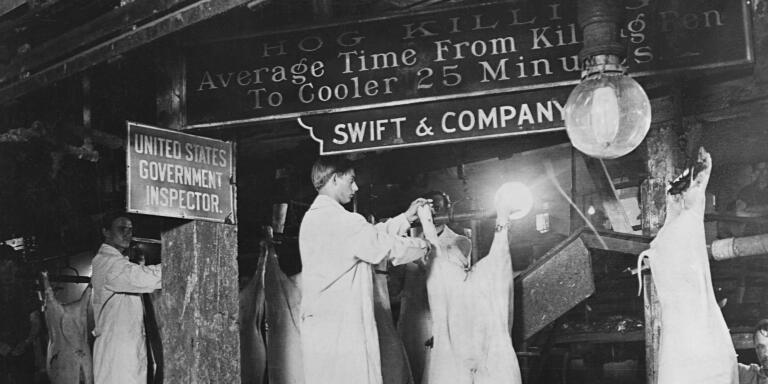 Meat inspectors examine hogs at the Swift & Company packing house in Chicago. The hogs are hanging, and the men wear long white coats. (Photo by © CORBIS/Corbis via Getty Images)