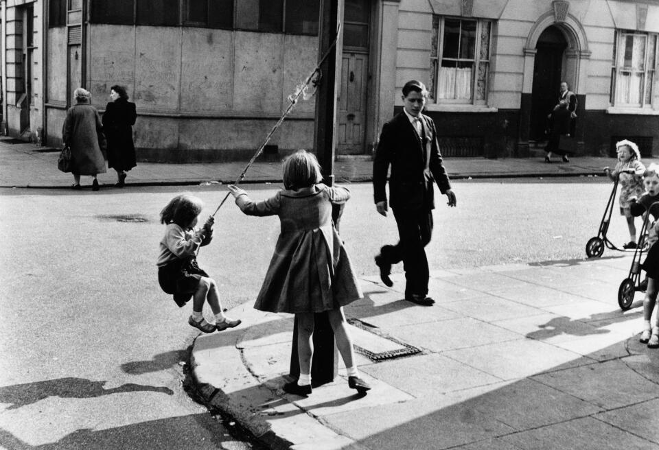Two girls playing on a street corner in North Kensington, London. The smaller girl swings from a rope attached to a lamppost. Other children are approaching on scooters, and a young man has just crossed the road. (1957)