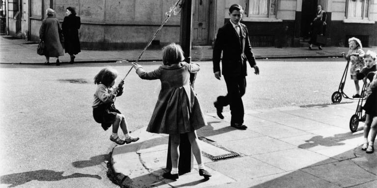 Two girls playing on a street corner in North Kensington, London. The smaller girl swings from a rope attached to a lamppost. Other children are approaching on scooters, and a young man has just crossed the road. (1957)
