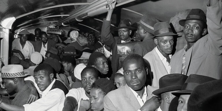 SOUTH AFRICA. 1960s. Africans throng Johannesburg station platform during late afternoon rush hour. Train accelerates with its load of clinging passengers. They ride like this through rain and cold, some for the entire journey. Inside, hands cling to a suitcase.