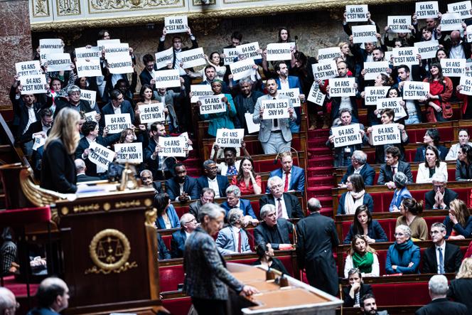 Des députés « insoumis » ainsi que d’autres élus de la Nupes brandissent des pancartes « 64 ans c’est non », lors du discours d’Elisabeth Borne, qui recourt à l’article 49.3 à l’Assemblée, afin de faire adopter la réforme des retraites sans vote de l’Hémicycle, le 16 mars 2023.