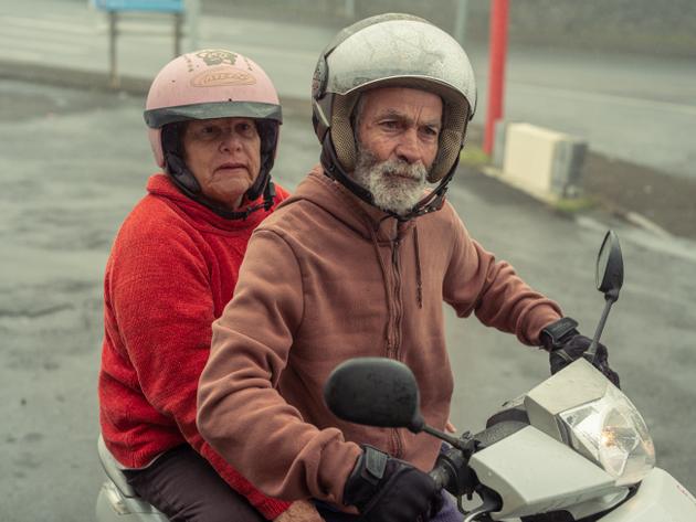 Jean-René et Suzie sortent de la Supérette du Volcan, où ils sont venus faire leurs courses à scooter du village voisin. Une fois le coffre rempli sous la selle, les derniers achats tiendront entre les jambes du conducteur. Suzie écoute son mari parler en levant les yeux au ciel. Le Tampon (La Réunion), le 18 juin 2024.