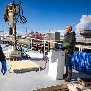 La Turballe, France - 17 avril 2025. Anthony Le Huche, marin-pêcheur et patron du chalutier le Magayant manipule une grue pour décharger des caisses de poisson à quai après une campagne de pêche tandis que son fils Théo Le Huche, 28 ans inspecte le pont du bateau. Photographie par Jérémie Lusseau / Hans Lucas pour Le Monde