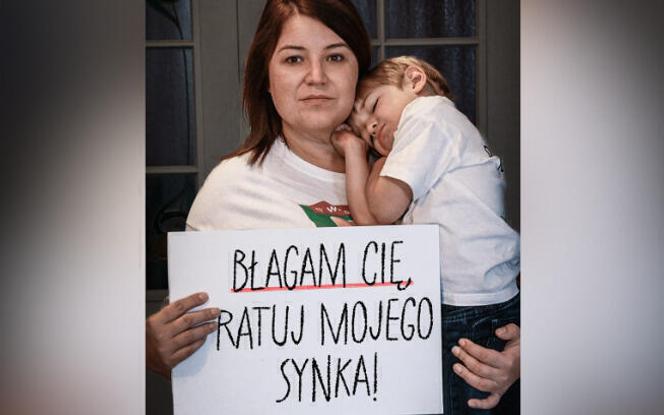 Dans une photo tirée du site polonais de cagnottes en ligne Siepomaga, Monika tient dans ses bras son enfant, Ignas, et une pancarte où l’on peut lire « Je vous supplie de sauver mon fils ».