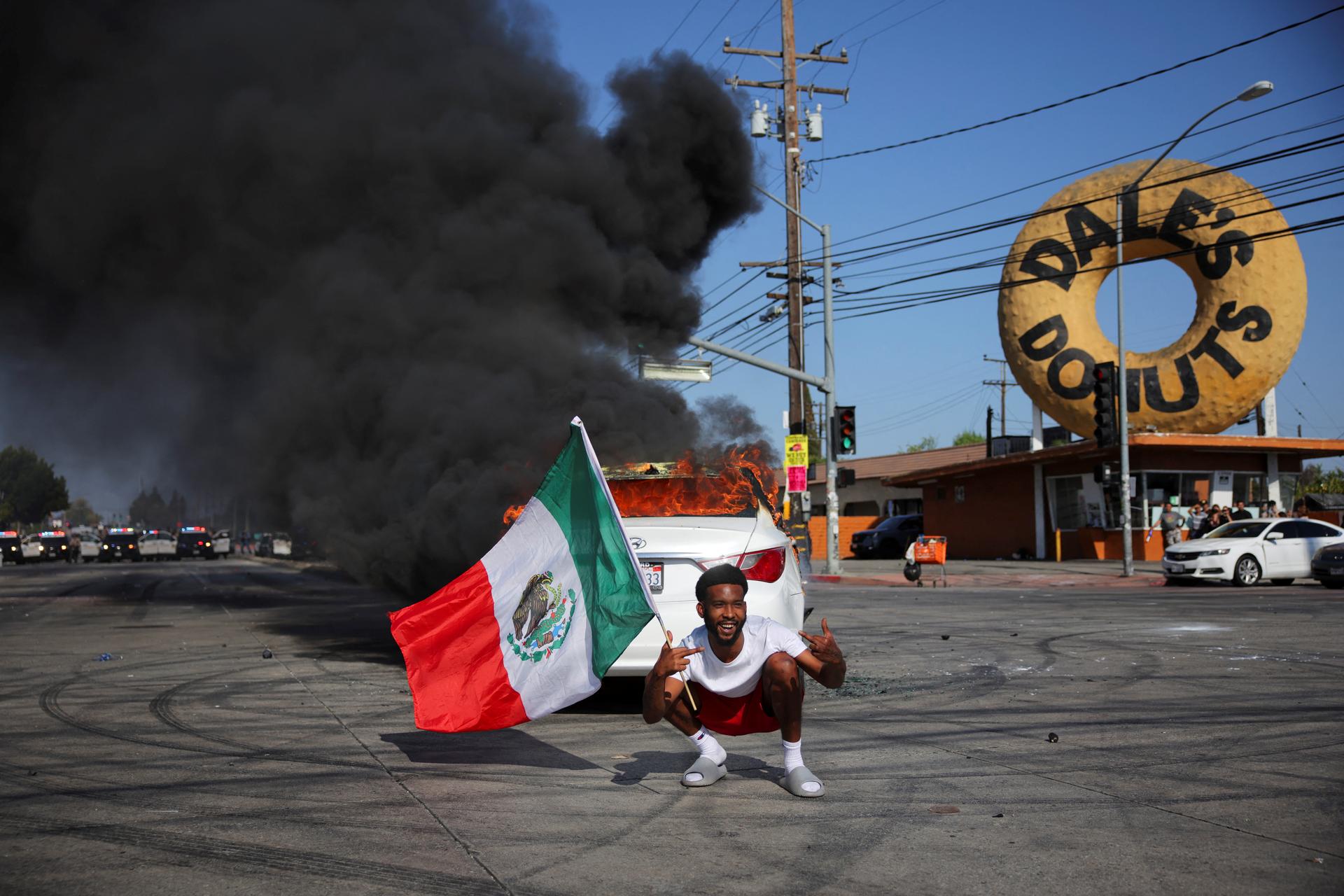 Un homme, drapeau mexicain en main, devant une voiture en feu, dans la ville de Paramount, comté de Los Angeles, Californie, le 7 juin 2025.