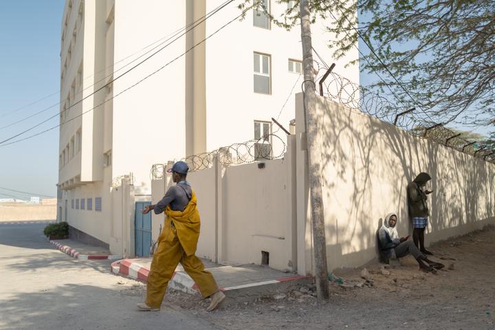 Un migrant en tenue de pêcheur se dirige vers le port artisanal. A droite, des migrants profitent du signal Wi-Fi d’un hôtel voisin pour surfer sur Internet, à Nouadhibou (Mauritanie), le 15 juin 2025.
