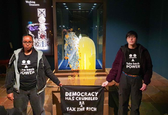 Des activistes du groupe de résistance civile Take Back Power brandissant une banderole sur laquelle est écrite « La démocratie s’est effondrée – Taxez les riches », après avoir jeté de la nourriture sur la vitrine protégeant les joyaux de la couronne d’Angleterre, à la tour de Londres, le 6 décembre 2025. 