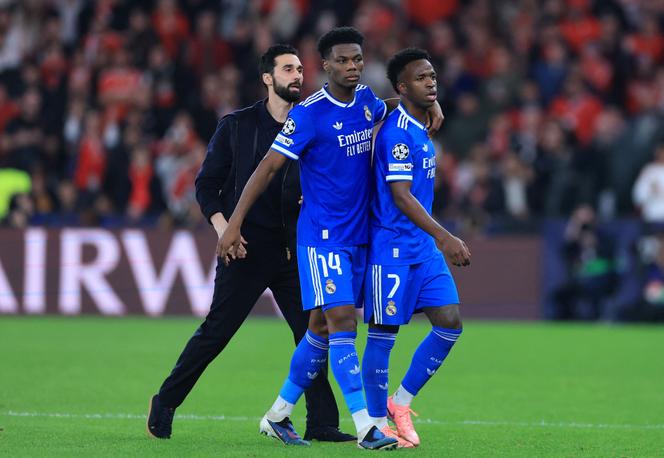 Vinicius (à direita), rodeado pelo seu treinador Álvaro Arbeloa e pelo seu companheiro de equipa Aurélien Tchouaméni, durante o jogo da Liga dos Campeões do Real Madrid no relvado do Benfica, a 17 de fevereiro de 2026.