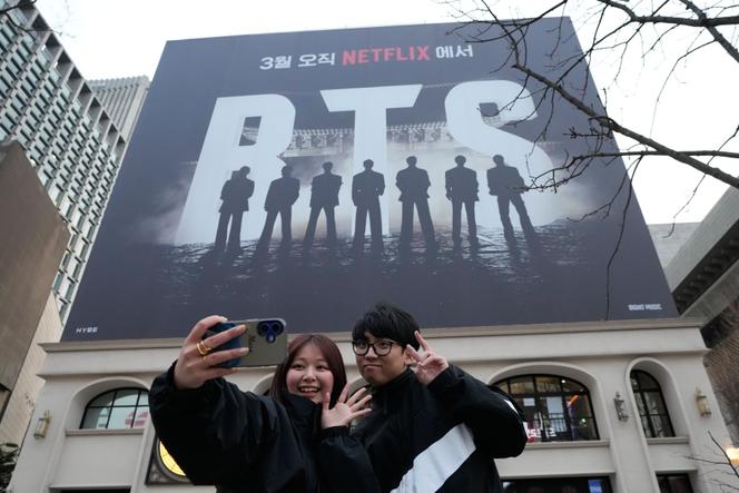 Un couple prend un selfie devant une affiche annonçant le concert de retour du groupe de K-pop BTS, sur la place Gwanghwamun, à Séoul, le 18 mars 2026.