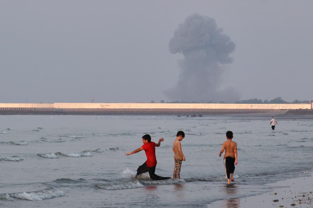 Trois enfants jouent dans les eaux du détroit d’Ormuz, alors qu’un panache de fumée s’élève de Bandar-e Abbas, en Iran, le 13 juillet 2026. - RAZIEH POUDAT/AP PHOTO
